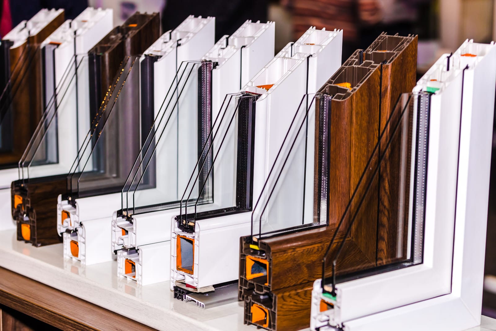Cutaway sections of window frames lined up on a sill, showing the hollow chambered interiors of white and wood-finish profiles with double- and triple-glazed units fitted